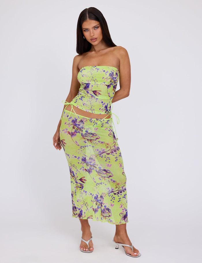 A person strikes a pose against a plain background donning the TIGERMIST Kellan Skirt - Green by Poppy Floral, featuring an abstract, tie-dye pattern in green, purple, and yellow shades. They pair this stylish piece with strappy white high-heeled sandals. The individual has long, dark hair.