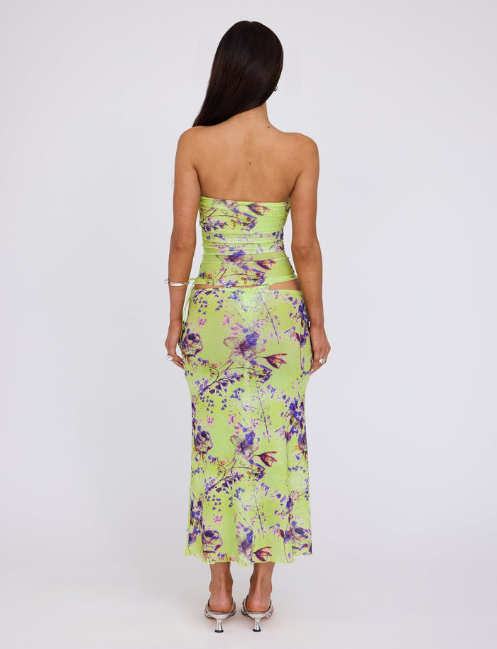 A woman with long dark hair wearing a strapless, green floral dress from TIGERMIST's KELLAN SKIRT - GREEN : BY POPPY FLORAL stands facing away from the camera. She is also wearing clear high-heeled shoes and a bracelet on her left wrist. The background is plain white.