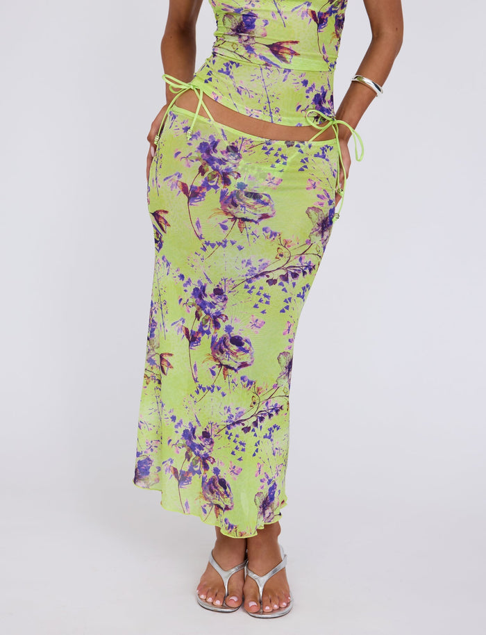 A person is wearing the KELLAN SKIRT - GREEN : BY POPPY FLORAL from TIGERMIST, which is a light green maxi skirt with a purple and pink floral print. The skirt features side cutouts with string ties. They are also wearing a matching top and white sandals. Only their torso and legs are visible against a plain white background.