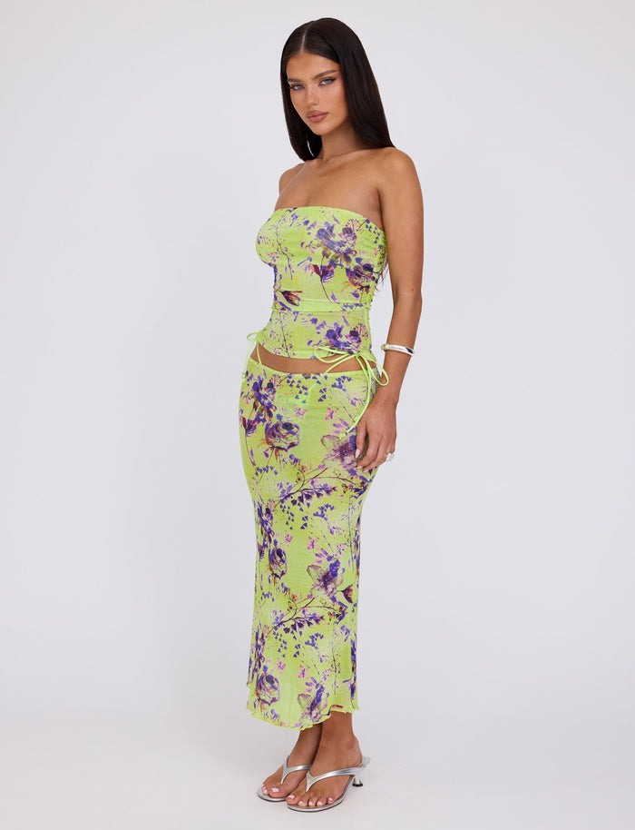 A woman with long dark hair stands against a plain white background. She is wearing the KELLAN SKIRT - GREEN by Poppy Floral from TIGERMIST, which features a strapless, form-fitting design in lime green adorned with a purple floral print and cut-out details on the sides. She completes her outfit with white open-toed heels and a bracelet.