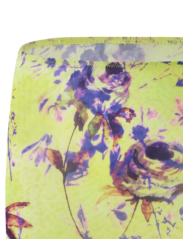 A close-up of the KELLAN SKIRT - GREEN from TIGERMIST, featuring an abstract floral design by Poppy Floral. The background is green, adorned with flowers in shades of purple and light pink. The stitching on the top edge is visible, and the fabric appears to be translucent with a soft, lightweight texture.