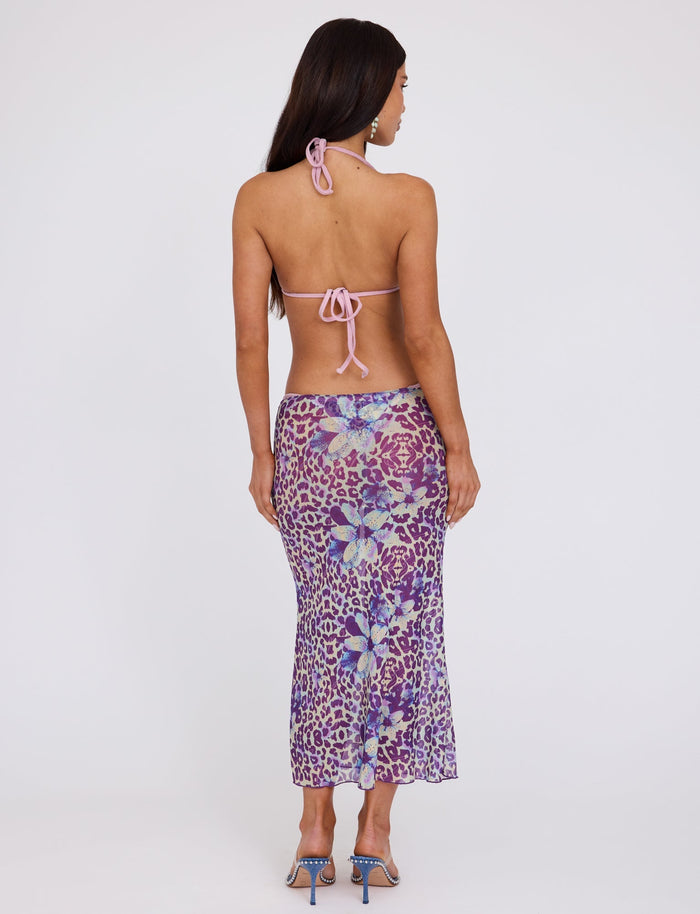 A woman stands with her back to the camera, showcasing a light pink backless halter top and the KELLAN SKIRT - PURPLE : BY POPPY LEOPARD from TIGERMIST, an ankle-length skirt featuring a vibrant floral and leopard print pattern. Her dark hair cascades down as she steps confidently in blue high-heeled shoes.