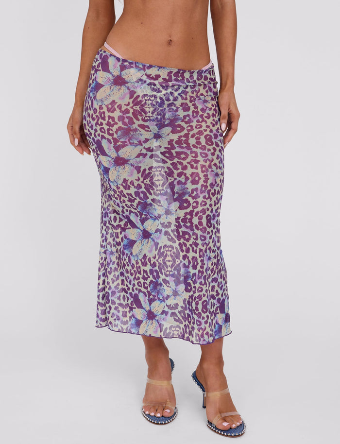 A person modeling a sheer, high-waisted Kellan Skirt-Purple by Poppy Leopard from TigerMist, featuring a purple leopard and blue floral print pattern. The skirt is paired with clear high-heeled sandals against a plain white studio backdrop.