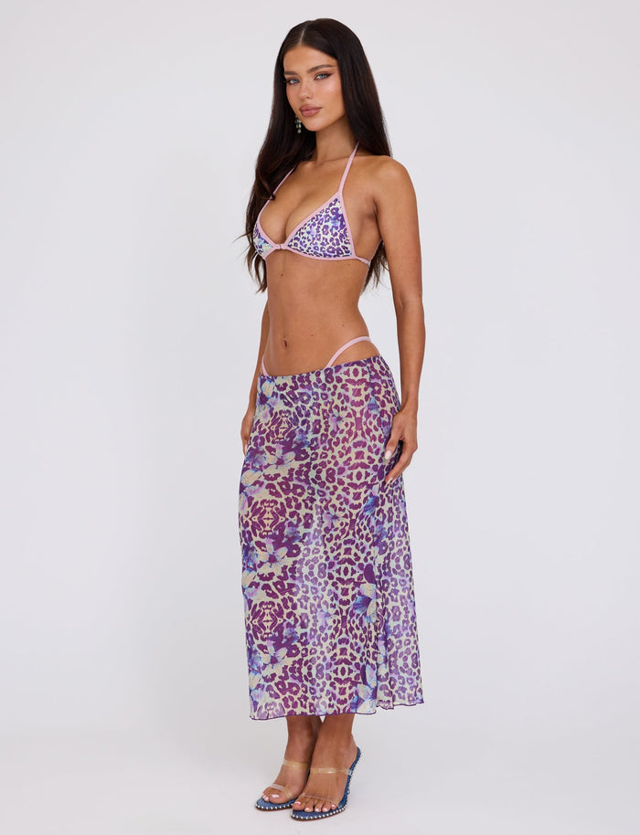 A person with long dark hair is wearing a Tiger Mist purple leopard print bikini top and the KELLAN SKIRT - PURPLE: BY POPPY LEOPARD, a matching sheer high-waist skirt from TIGERMIST. They are also wearing blue high-heeled sandals and standing against a plain white background, looking slightly to the side.