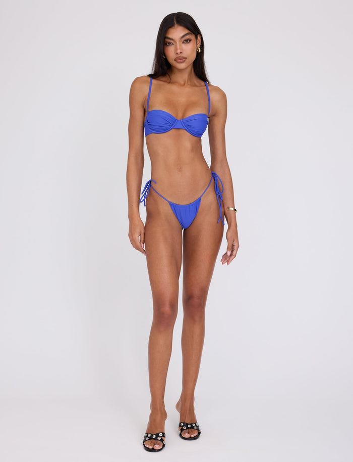 A woman stands against a plain white background wearing the chic KHESSA BIKINI BOTTOM in cobalt blue from TIGERMIST. She has long dark hair and is posing with a neutral expression. Additionally, she is wearing black sandals and has a bracelet on her left wrist.