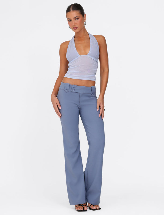 A person poses confidently against a white background, wearing a sleeveless, halter-neck lavender top paired with the KITTIE PANT in steel blue by TIGERMIST. They accessorize with hoop earrings and black heeled sandals.