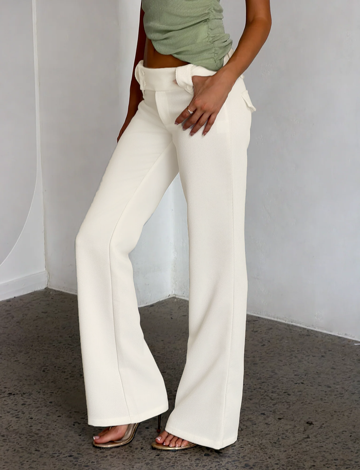 A person stands indoors on a textured gray floor, wearing TIGERMIST's KITTIE PANT - WHITE : CREAM with a flare fit and a light green sleeveless top, one hand resting in their pocket.