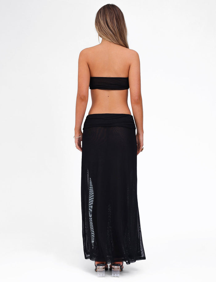 A woman is standing with her back to the camera, wearing the black KYNLEE MAXI DRESS by TIGERMIST. She has long, light brown hair and is paired the dress with high-heeled shoes. The background is plain white.