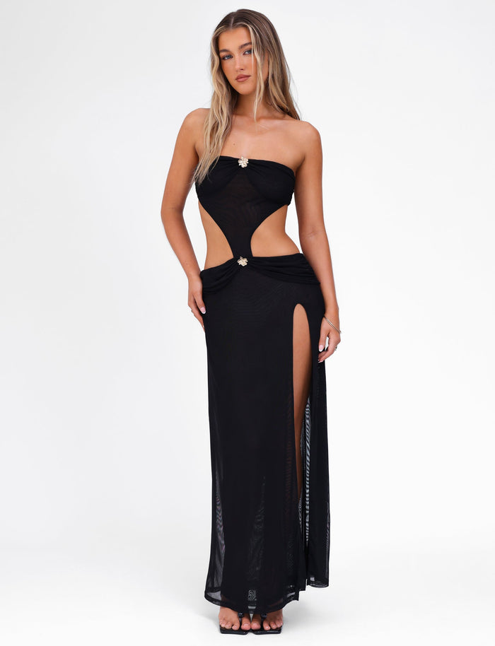 A person is wearing the TIGERMIST KYNLEE MAXI DRESS - BLACK, a strapless black evening gown featuring waist cutouts and a high slit on one side, embellished with floral gold metal hardware. Their long, wavy blonde hair cascades over their shoulders as they stand against a plain white background.