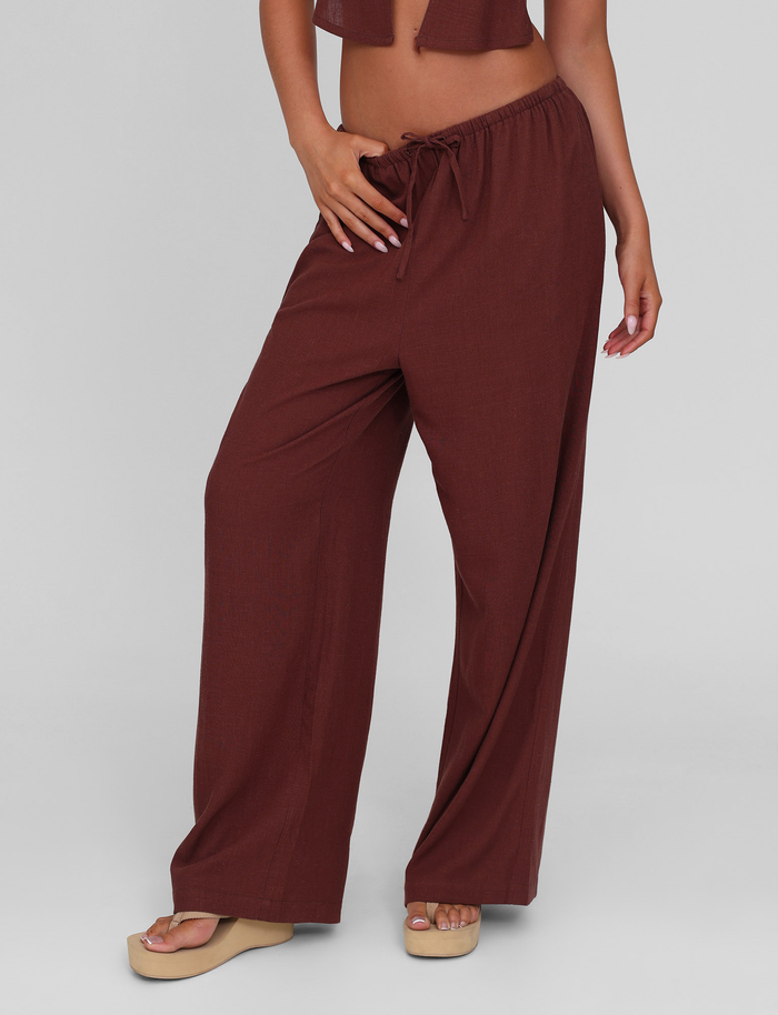 A person models the TIGERMIST LAINEY PANT in chocolate, featuring a loose fit, wide legs, and an elasticated drawstring waist. Paired with beige platform sandals, the cropped image is set against a plain light grey background.