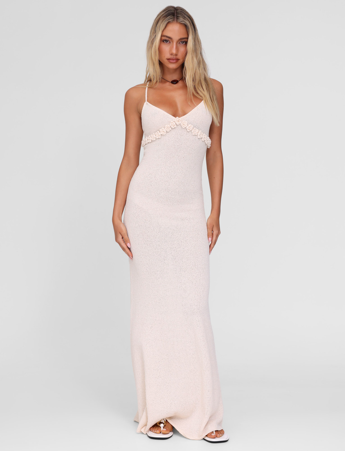 A woman poses against a plain background in the TIGERMIST LAVELLE MAXI DRESS in cream, featuring thin crisscross straps and a floral chest accent. She has long, loose blonde hair and neutral makeup.