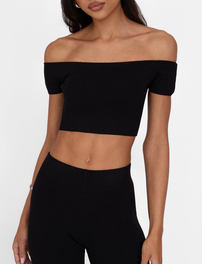 A woman is wearing the sleek LEDGER TOP - BLACK by TIGERMIST, an off-shoulder crop top with short sleeves that highlights her midriff. Paired with high-waisted pants creating a fitted silhouette, she stands against a plain white background, her long dark hair cascading down.