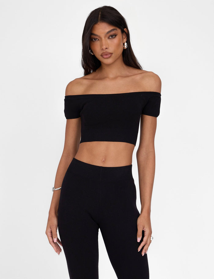 A person with long, dark hair is wearing the LEDGER TOP - BLACK from TIGERMIST, along with high-waisted black pants, creating a fitted silhouette. They are standing against a plain white background, looking directly at the camera with a neutral expression. They accessorize their outfit with earrings and a bracelet.