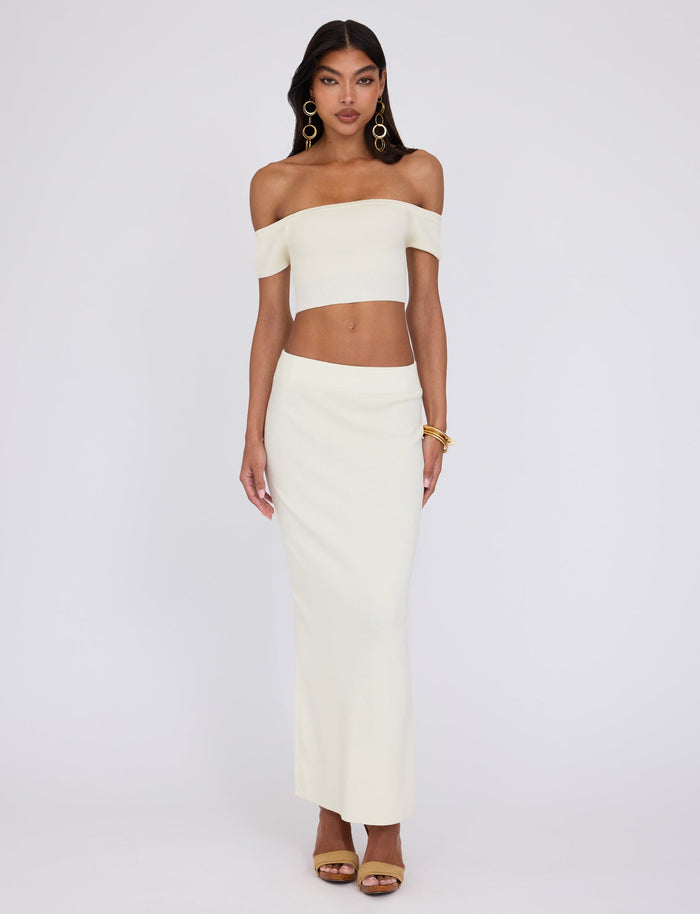 A woman stands against a plain white background, showcasing a chic outfit by TIGERMIST. She wears the LEDGER TOP - WHITE : CREAM, an off-the-shoulder, cream-colored crop top, paired with a matching floor-length skirt. With long dark hair, gold hoop earrings, gold bangles on her left wrist, and beige high-heeled sandals, she exudes effortless elegance.