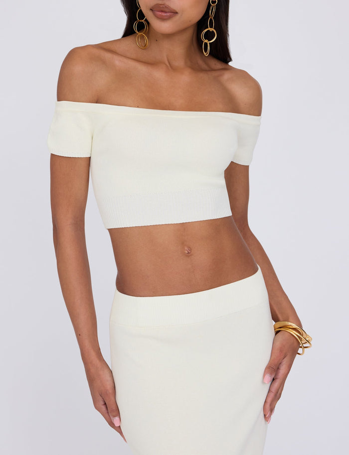 A person is wearing a light-colored off-the-shoulder crop top named "LEDGER TOP - WHITE : CREAM" from TIGERMIST and a matching high-waisted skirt. They have long, straight hair and are accessorized with large hoop earrings and gold bangles on their left wrist. The background is plain white.