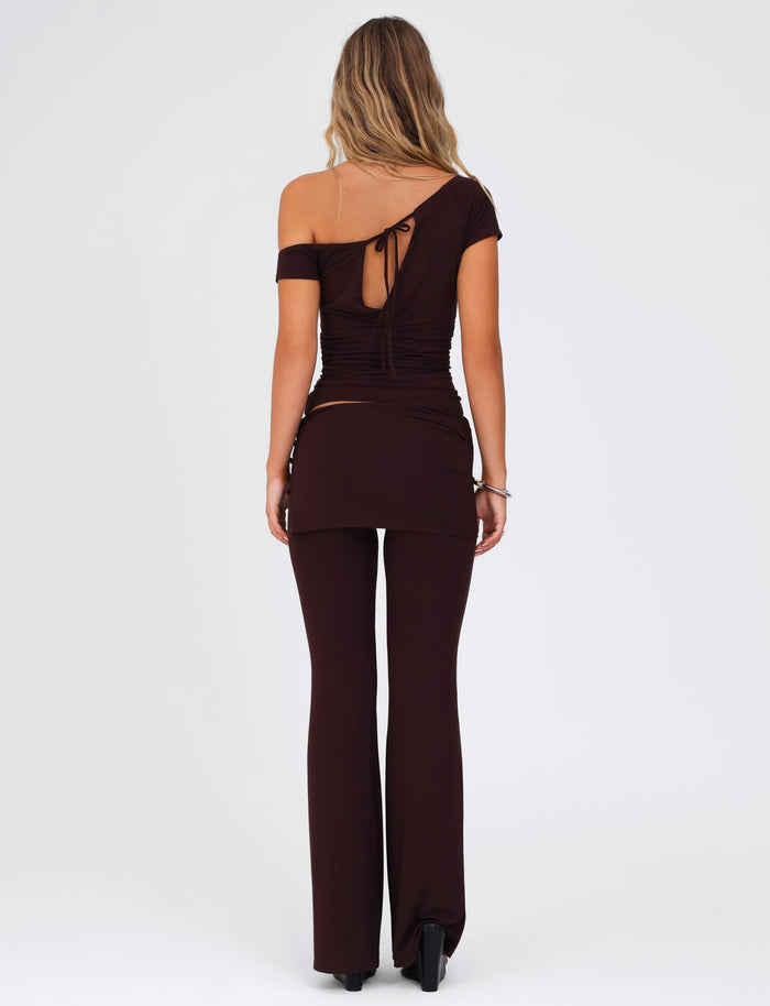 A woman stands with her back turned, wearing the TIGERMIST LEILANI PANT in a chocolate brown hue, accompanied by a chic off-shoulder top featuring a tie at the back and made from high-stretch fabric. Her long, wavy hair flows over the coordinated ensemble against a simple, light background.