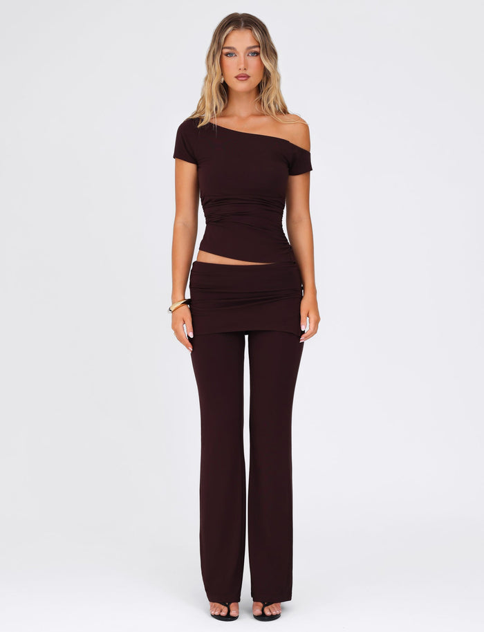 A person stands against a plain background, wearing the LEILANI TOP in chocolate by TIGERMIST, featuring a stylish asymmetrical design and one-shoulder neckline. They pair it with wide-leg pants made from buttery jersey and accessorize with a gold bracelet and black heels.
