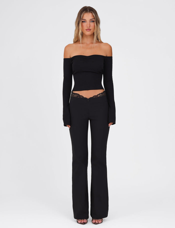 A woman stands against a white background wearing the TIGERMIST LILYANNA LONG SLEEVE TOP in black, paired with high-waisted lace-detailed pants. Her long, wavy blonde hair cascades over her shoulders as she completes the look with open-toed heels.