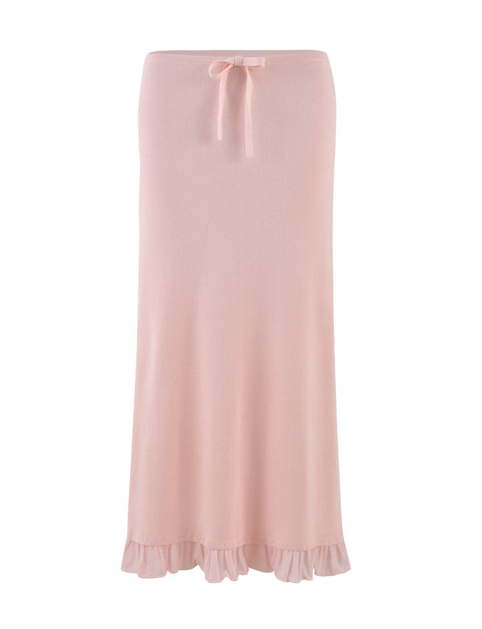 The TIGERMIST LILYANA SKIRT in Baby Pink is a long, pastel pink knit skirt with a relaxed fit, featuring a ruffled hem and a small bow at the waist. This elegant midi skirt is set against a white background.