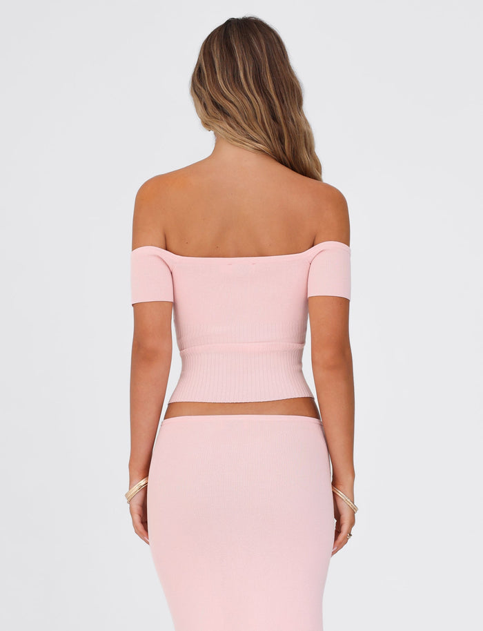 A woman with long hair is wearing the TIGERMIST LILYANNA TOP in baby pink, featuring an off-the-shoulder design and a matching stretchy skirt. The image captures her from the back against a simple white background.