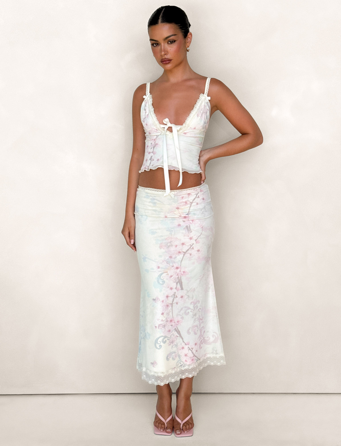 A woman with dark hair pulled back wears the TIGERMIST LONIE MIDI SKIRT in Washed Cherry Blossom Floral with a matching sleeveless bow top, standing against a plain light background and paired with heeled sandals.