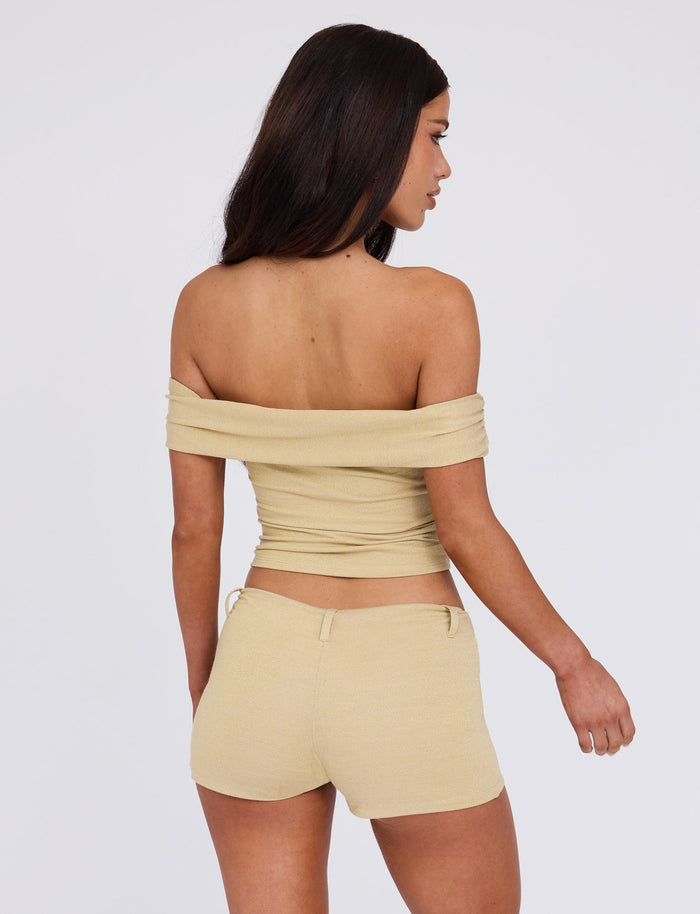 A woman with long dark hair faces away, wearing a beige off-the-shoulder top and matching high-waisted LYNSEY SHORT - GOLD from TIGERMIST. The background is plain white, emphasizing her outfit. She appears to be standing in a relaxed pose with her right arm slightly bent.