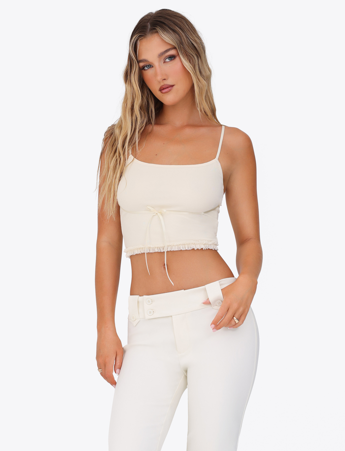 A woman with long, wavy blonde hair wears the TIGERMIST MAISIE TOP in pale yellow, featuring adjustable straps, paired with high-waisted white pants against a plain white background.
