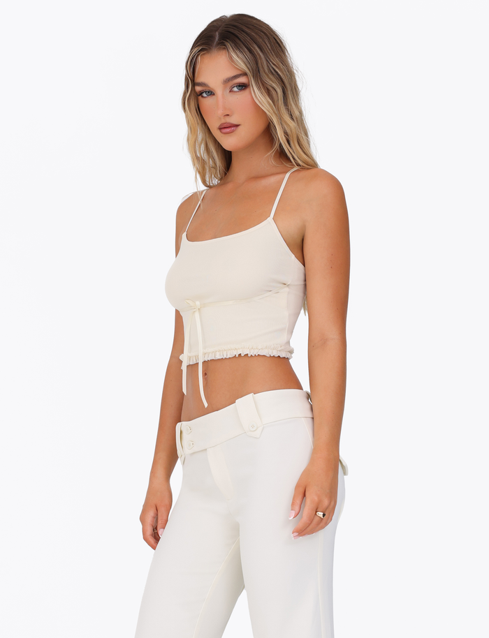 A woman with long, wavy blonde hair wears the TIGERMIST MAISIE TOP in pale yellow, standing against a plain white background and looking toward the camera.