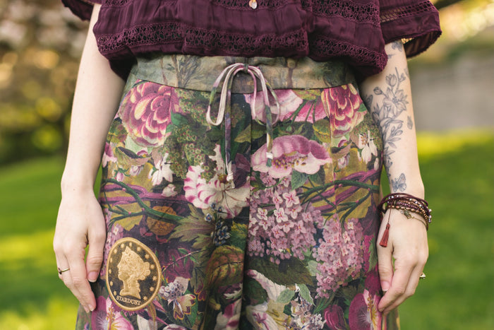 Model wearing linen blend artist pants featuring antique wishbone–Victorian coin–evening star–dandelion art and “Wish with Me” banner on floral background – Market of Stars