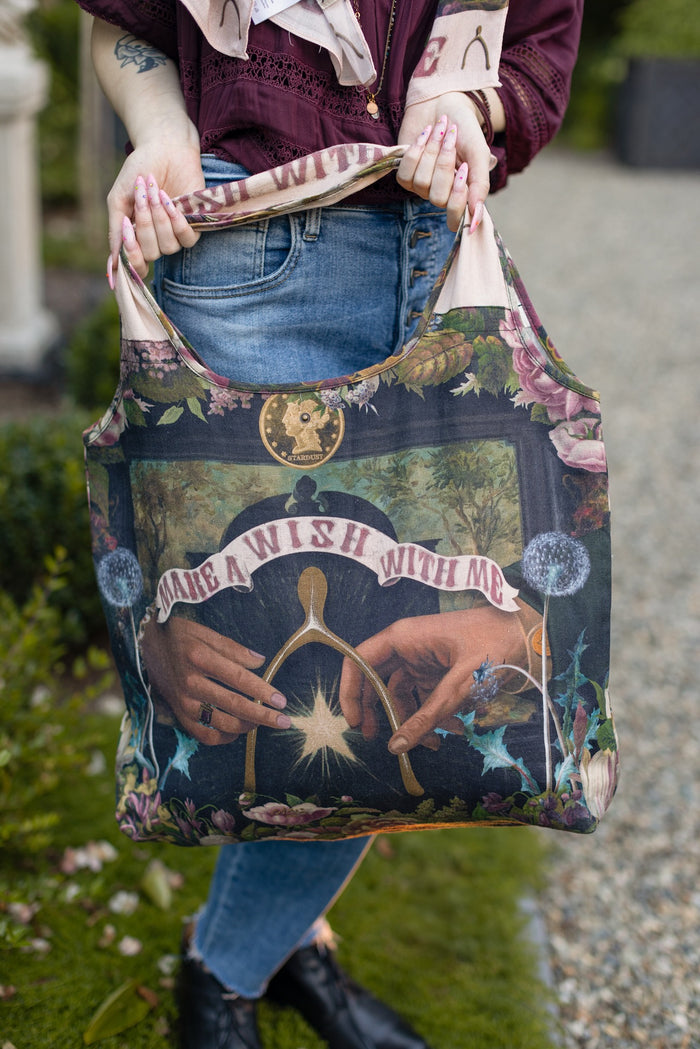 Model holding linen blend market tote featuring Victorian wishbone–vintage coin–evening star–dandelion–and “Best Wishes” banner on floral background – Market of Stars