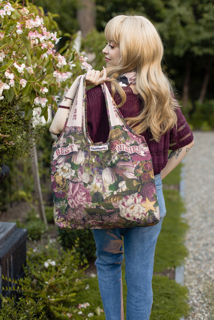 Model holding linen blend market tote featuring Victorian wishbone–vintage coin–evening star–dandelion–and “Best Wishes” banner on floral background – Market of Stars