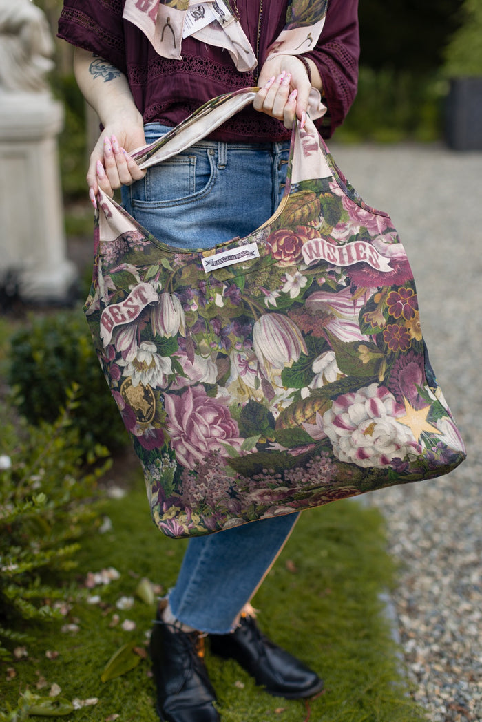 Model holding linen blend market tote featuring Victorian wishbone–vintage coin–evening star–dandelion–and “Best Wishes” banner on floral background – Market of Stars