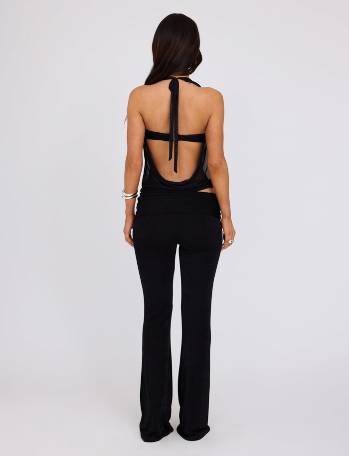 A person with long dark hair is standing with their back to the camera against a plain white background. They are wearing a black, backless halter top and TIGERMIST's MAKENA PANT in black. The top is tied at the neck with a strap, and they have a bracelet on their left wrist.