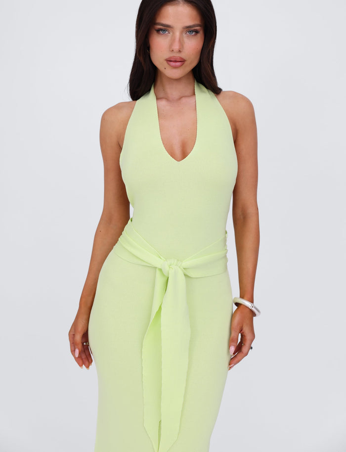 A woman with long brown hair is wearing the MAREE DRESS - GREEN by TIGERMIST, a light green knit dress in a halter style with a knotted belt at the waist. She stands against a plain white background, one arm resting by her side and the other slightly bent. A silver bracelet adorns her right wrist.