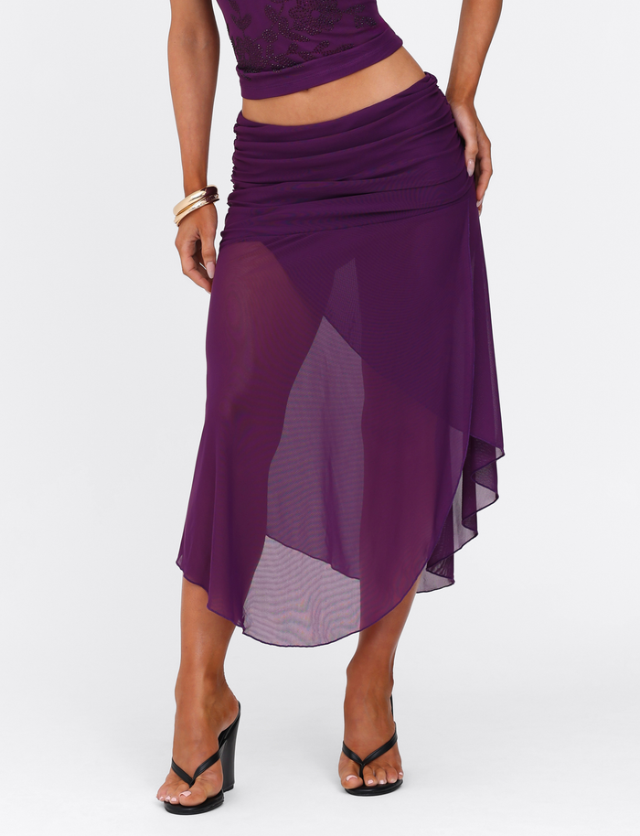 A person stands against a plain white background wearing the TIGERMIST MARIANA SKIRT - PURPLE, styled with its matching top, high-heeled black sandals, and gold bangles.