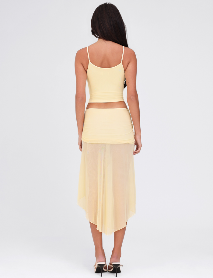 A woman with long dark hair, seen from behind, wears the TIGERMIST MARIANA SKIRT in pale yellow with a matching sleeveless crop top and high-heeled sandals, standing against a plain white background.