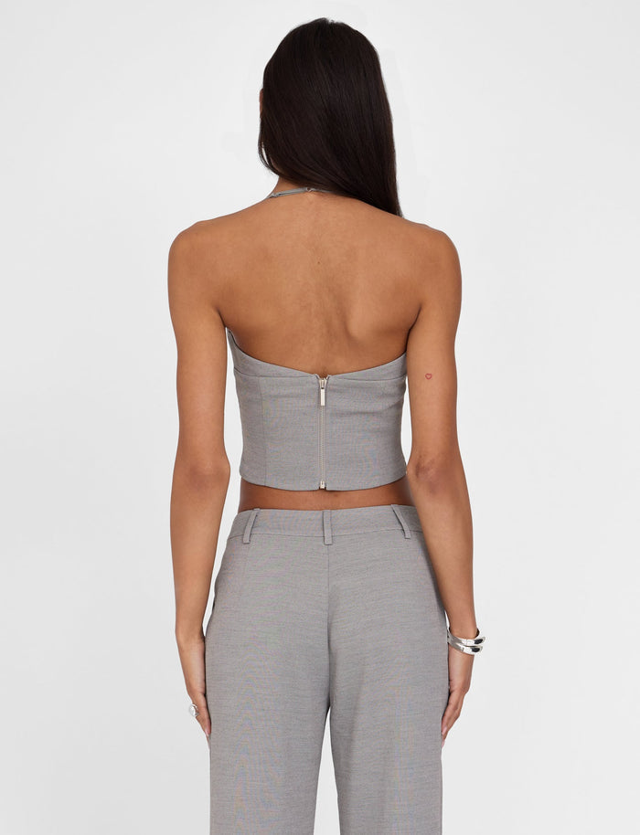 A woman with long dark hair is seen from the back, wearing the TIGERMIST MCKAY TOP in grey, which features a strapless design and a tailored bodice. She pairs it with matching high-waisted pants. Several silver bracelets adorn her left wrist. The background is plain white.
