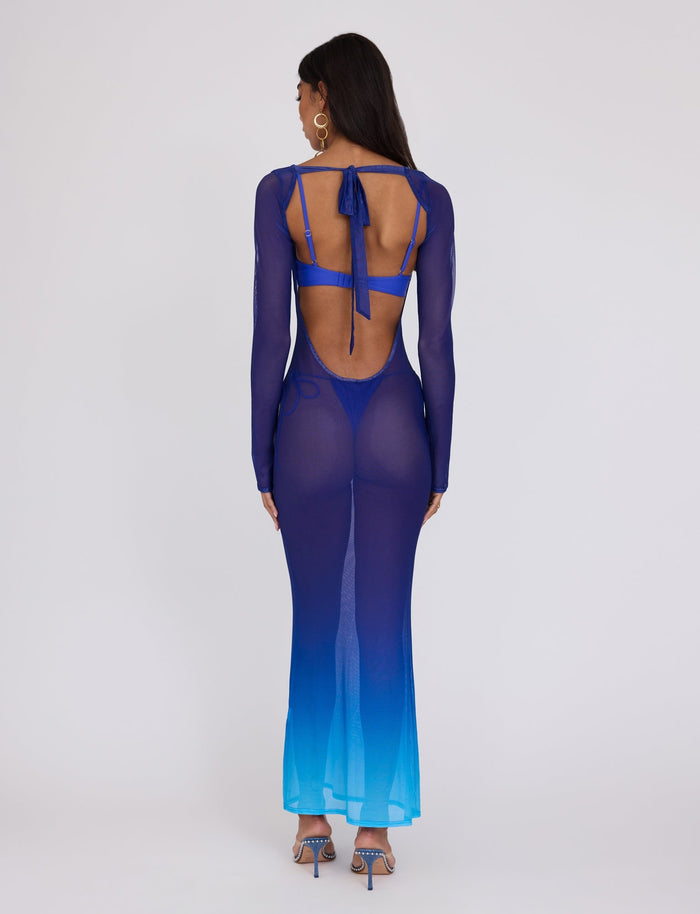 A woman stands with her back to the camera, wearing the MEILANI DRESS - BLUE : OMBRE from TIGERMIST. The long-sleeved, sheer dress features an open back and tie details at the shoulder blades. She has long, dark hair and is accessorized with strappy blue high-heeled shoes and large hoop earrings.