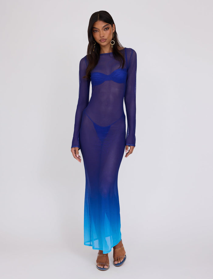 A woman with long, dark hair models the MEILANI DRESS - BLUE : OMBRE from TIGERMIST. The sheer maxi dress features a stunning gradient that transitions from dark blue at the top to light blue at the bottom. It has long sleeves and is paired with a bikini-style undergarment. She completes her look with silver hoop earrings.