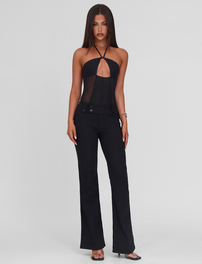 A woman with long straight hair wears the TIGERMIST MELANIA BODYSUIT in black, paired with high-waisted black pants. She stands facing forward against a plain white background, looking neutral with one arm by her side.