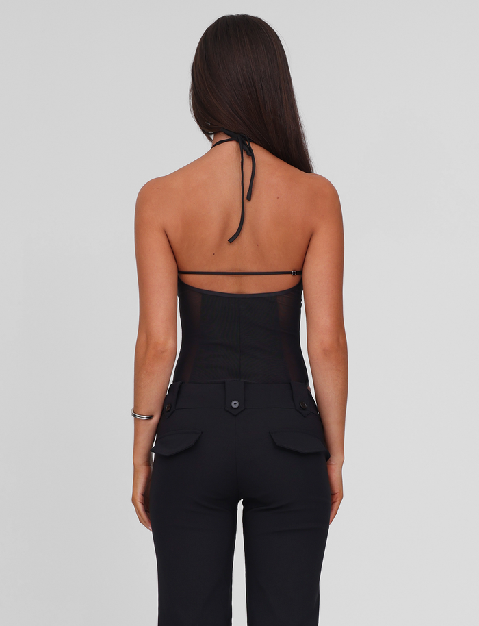 A woman with long brown hair faces away, wearing the TIGERMIST MELANIA BODYSUIT in black— a halter neck mesh style with an exposed back—paired with black pants featuring buttoned pockets, set against a plain light gray background.