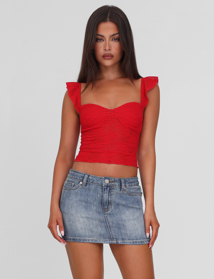 A woman with long dark hair models the TIGERMIST MICHAYLA MINI SKIRT - MID WASH, styled with a red lace crop top with ruffled sleeves, against a plain white background.