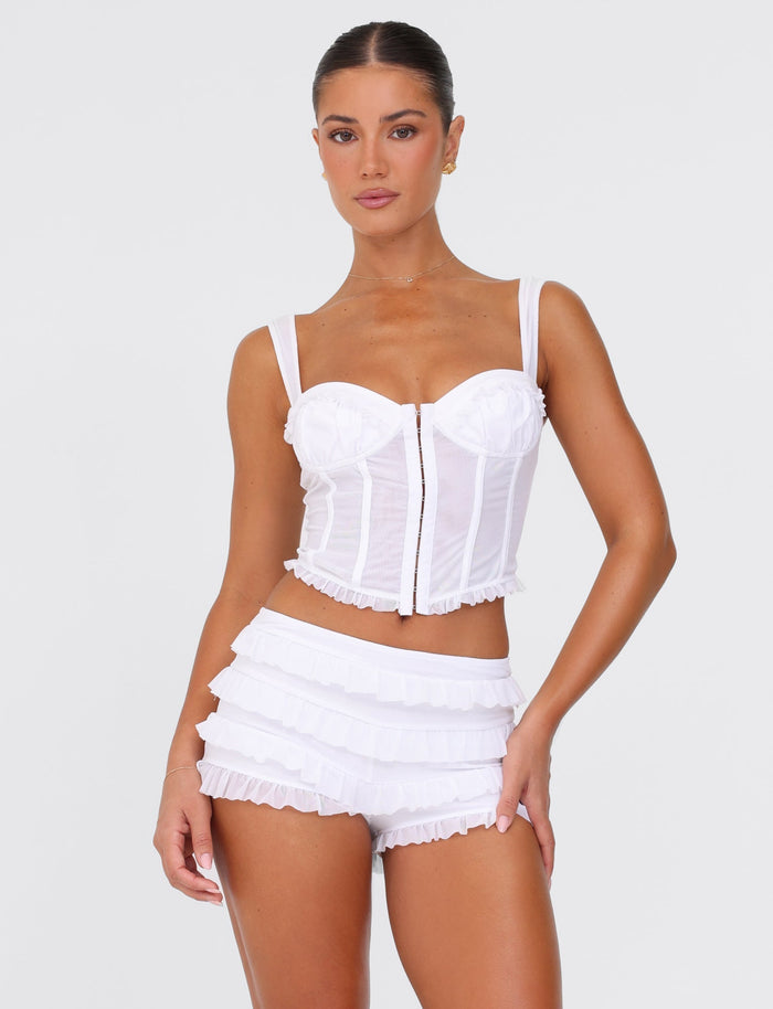 A woman stands confidently wearing the TIGERMIST MIKEL SHORT in white paired with a matching corset top. The outfit, showcasing a bodycon fit and ruffle details, exudes elegance through its delicate design. She poses effortlessly against a plain white background.
