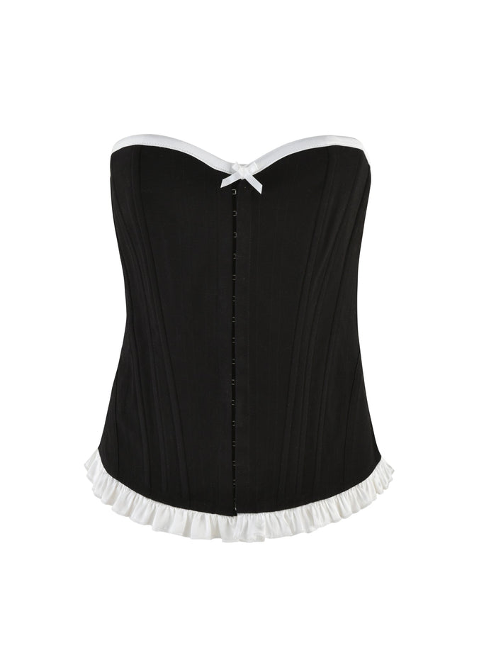 The MILLIE CORSET 2.0 - BLACK by TIGERMIST is a black denim corset featuring white trim along the top and bottom edges. This stylish piece has a white bow at the center of the neckline and a ruffled design at the hemline. Its vertical seams provide structure, while the lace-up back and front closure allow for versatile styling options.