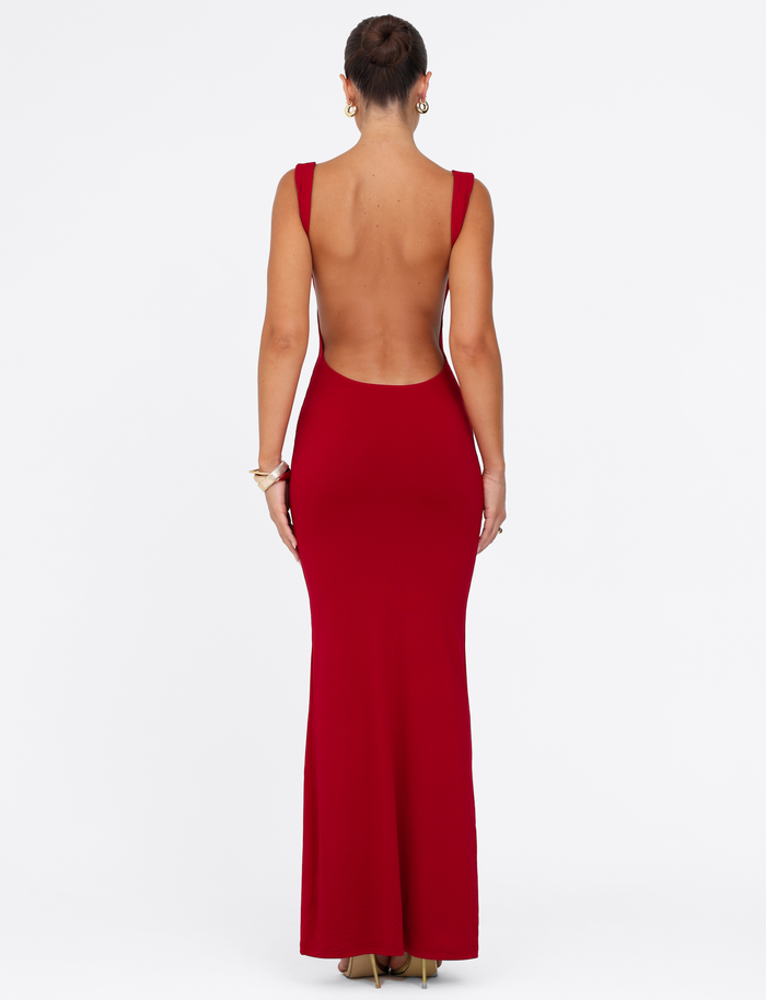 A woman faces away wearing the TIGERMIST MORANA MAXI DRESS - RED, a floor-length, sleeveless, backless bodycon dress paired with gold heels. Her hair is up, accessorized with earrings and bracelets, set against a plain white background.