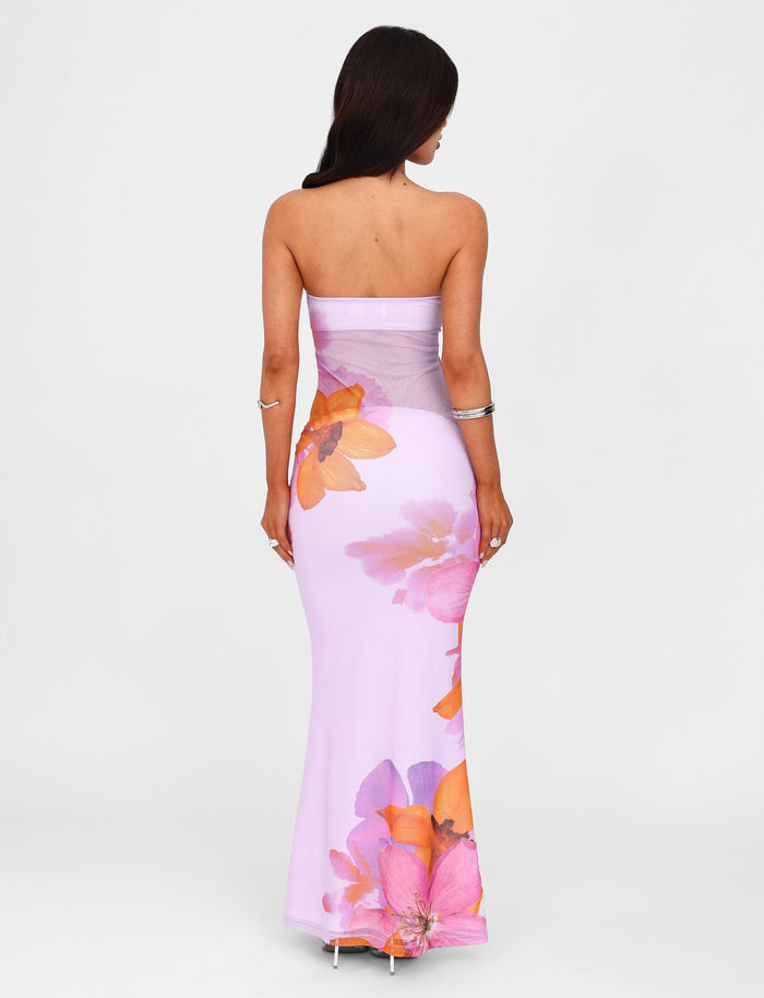 A woman with long brown hair is facing away, showcasing the MORGAN DRESS - PINK from the TIGERMIST By Poppy Summer collection. The strapless, floor-length lilac gown features large, vibrant orange and pink floral print designs. She completes her look with a silver bracelet against a plain white background.