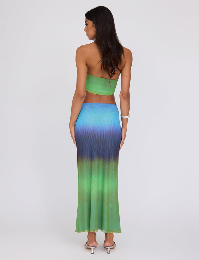 A woman stands with her back to the camera, showcasing a strapless green crop top paired with the NOELY SKIRT - MULTI : OMBRE : GRADIENT STRIPE from TigerMist. The skirt features a beautiful gradient transition from green at the waist to blue and purple hues towards the hem. She has long, dark hair and completes her outfit with white heels and a bracelet.