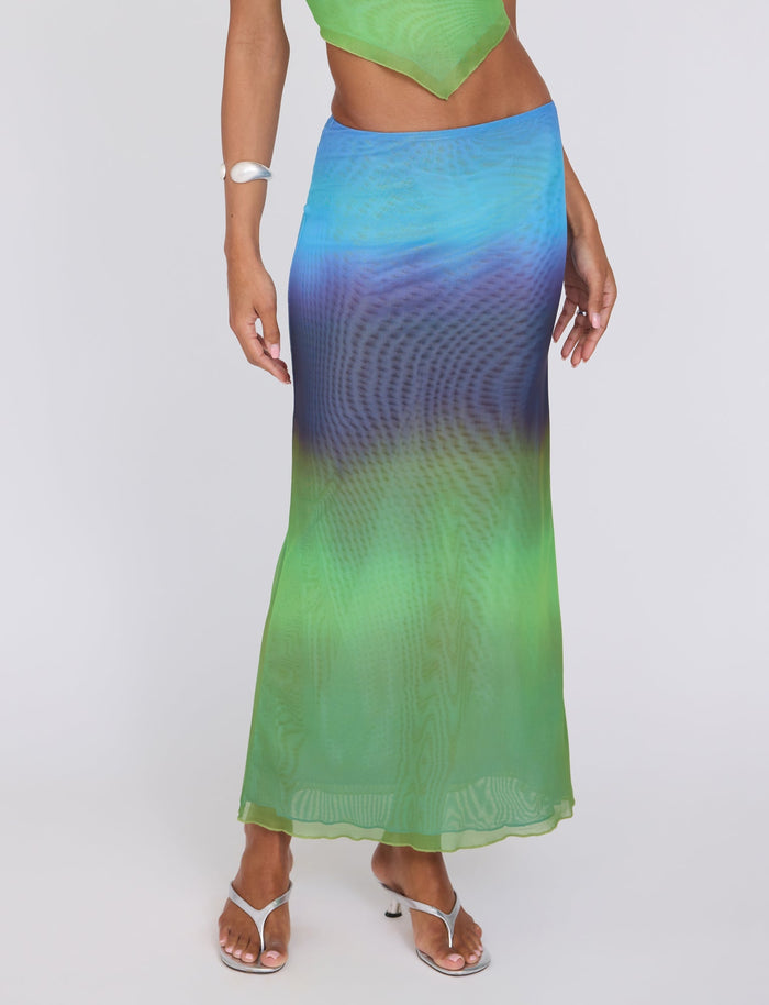 A person model is wearing the NOELY SKIRT - MULTI : OMBRE : GRADIENT STRIPE from TIGERMIST, featuring a bright and colorful gradient with hues of green, blue, and purple. They have paired it with a matching green top, a silver bangle, and white heeled sandals. The background is plain white.