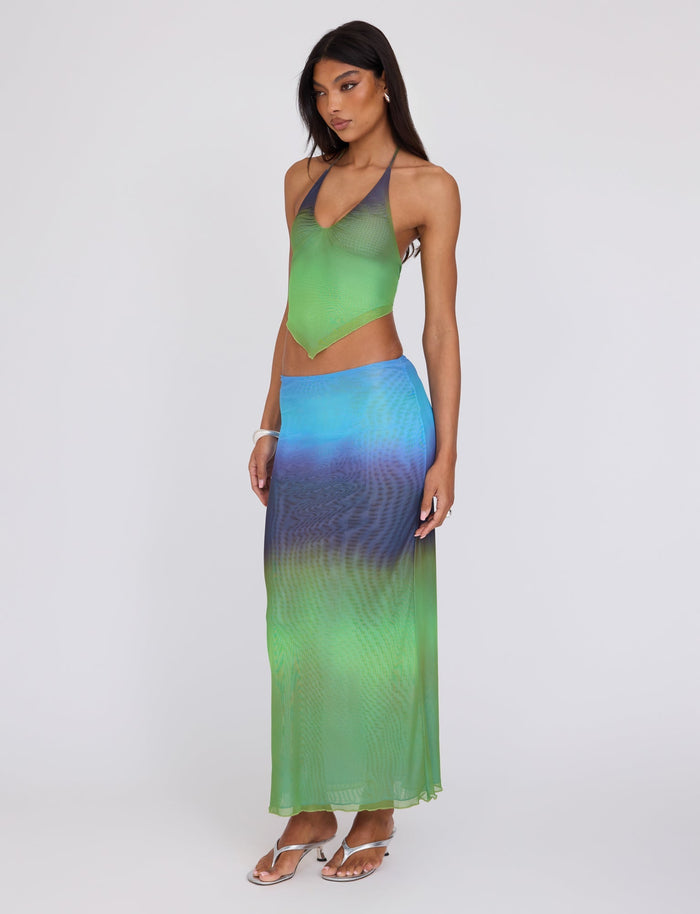 A woman stands against a plain background wearing the TIGERMIST NOELY SKIRT in a multi-colored ombre gradient stripe, with colors transitioning from green to blue to purple. She pairs it with a matching halter-neck top and accessorizes with silver sandals and a bracelet, her hair styled straight and loose.