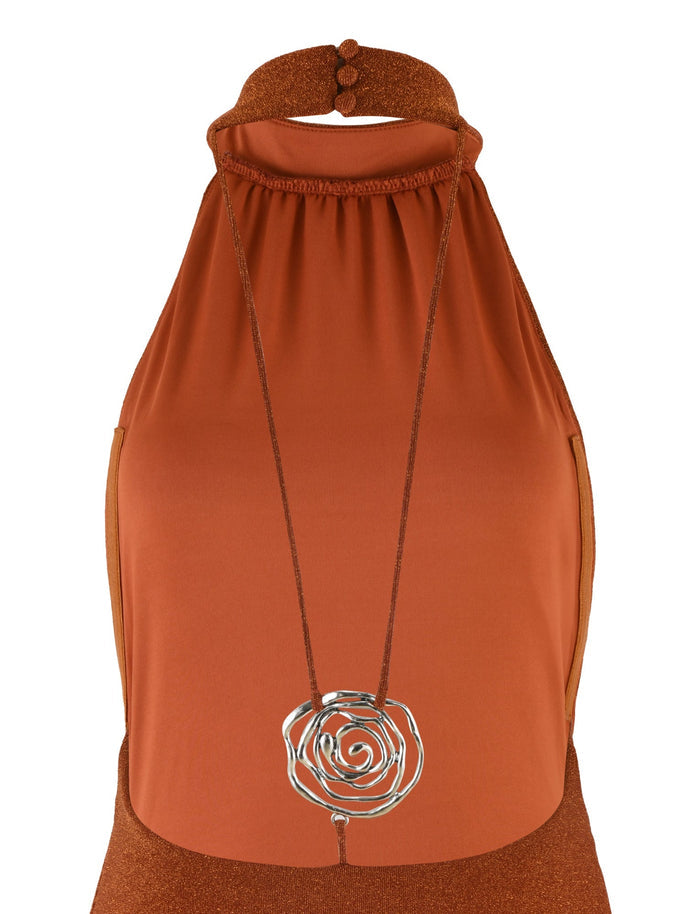 The NOVAK DRESS in orange by TIGERMIST is showcased from the chest up. It’s a sleeveless dress with a halter neckline and features a metallic, abstract flower pendant on a long cord around the neck. The shimmer knit fabric adds an elegant touch, with the neckline secured by two buttons at the back.